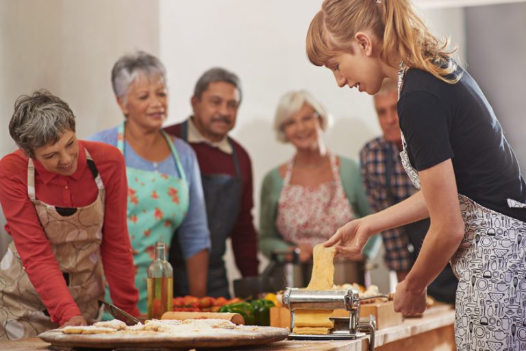 Food, senior cooking class and a woman teaching people in the kitchen of a home for meal preparation. Pasta maker, equipment and learning with mature friends watching a female chef follow a recipe