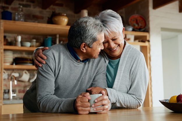 Happily retired elderly multi cultural couple smiling heads together wife holding husband in modern kitchen Happily retired elderly multi cultural couple smiling heads together wife holding husband in modern kitchen