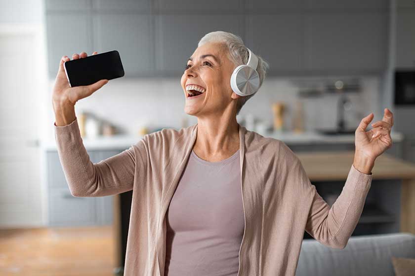 Cheerful European Senior Lady With Smartphone And Headphones Singing Song, Having Fun While Listening To Music At Modern Home Interior. Musical Mobile Application Concept