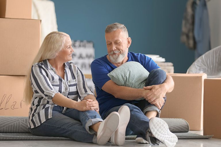 Mature couple with belongings sitting on floor indoors. Moving into new house