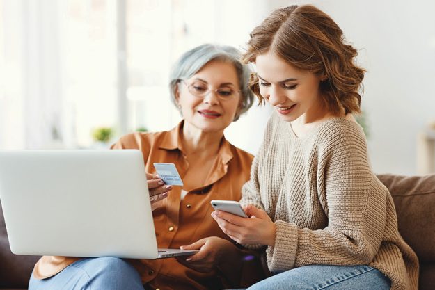 Positive young female with gray haired mother sitting on sofa with laptop and using smartphone while performing online transactions with credit card during shoppin