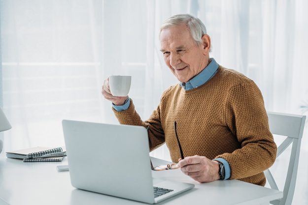 Senior confident man working on laptop and holding coffee cup