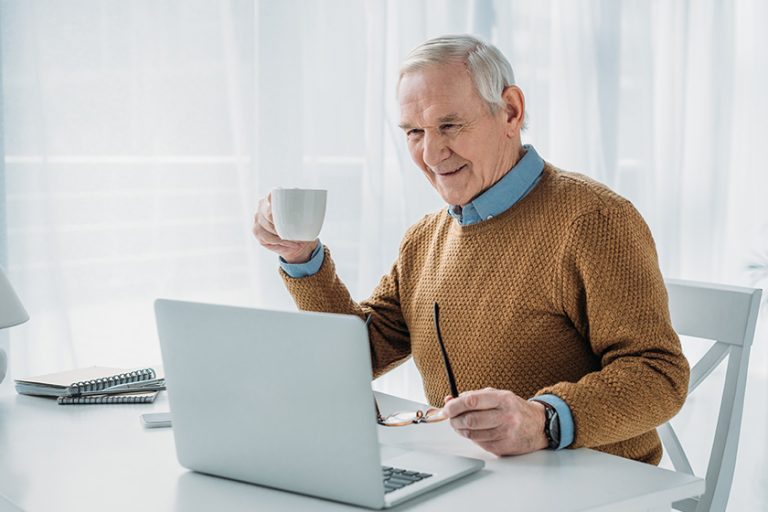 Senior confident man working on laptop and holding coffee cup