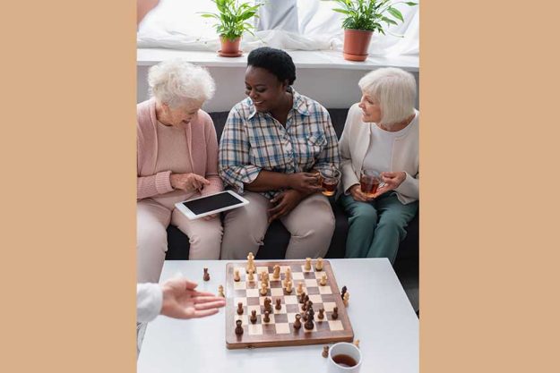 Senior woman pointing at digital tablet near interracial friends with tea and chess in nursing home Senior woman pointing at digital tablet near interracial friends with tea and chess in nursing home