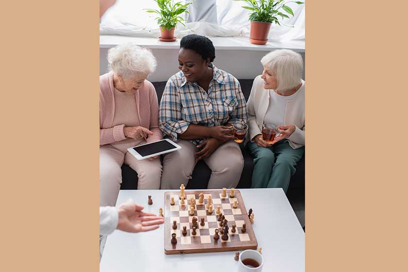 Senior woman pointing at digital tablet near interracial friends with tea and chess in nursing home Senior woman pointing at digital tablet near interracial friends with tea and chess in nursing home