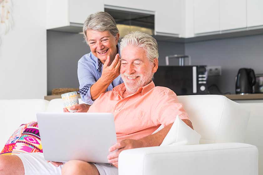 Aged couple sitting on sofa, drinking tea and using laptop at home Aged couple sitting on sofa, drinking tea and using laptop at home