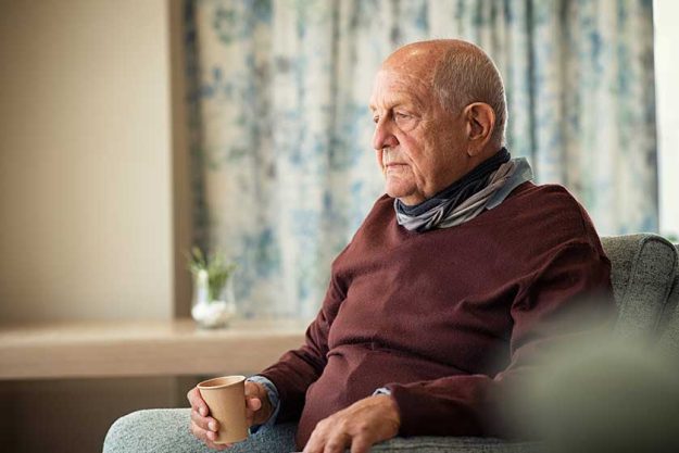 Depressed senior man sitting on armchair holding disposable cup of coffee and thinking. Frustrated retired man sitting on sofa and drinking a cup of hot tea. Sad mature man sitting alone at nursing home with sad expression.