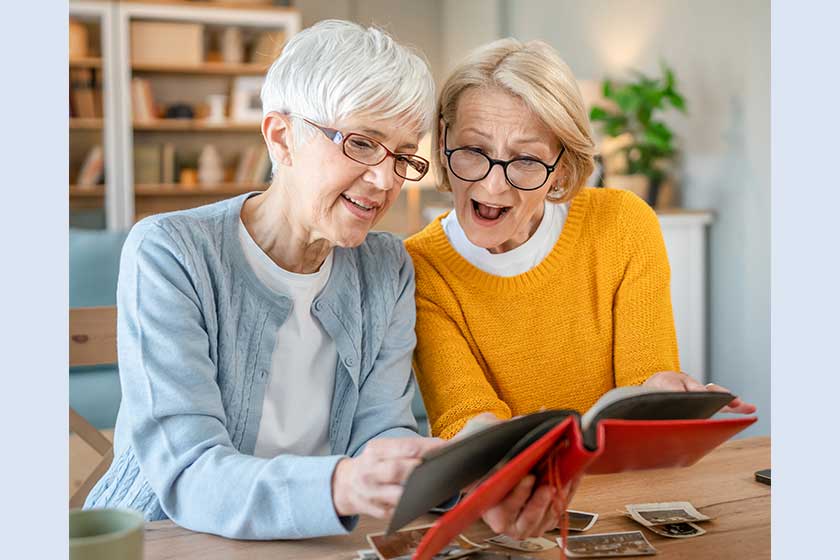 Senior mature women friends or sisters looking family photos album two caucasian females sit at home happy smile while revoke memories real people copy space