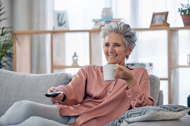 Senior woman, sofa and coffee to relax with remote for television, video or movies with smile in house. Elderly lady, couch and tea with happiness in home living room for watching tv in retirement.