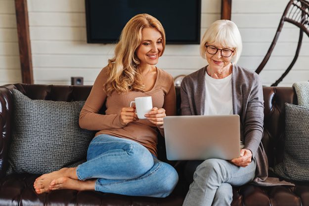 Smiling senior woman and her middle aged daughter sitting relax on sofa and drinking coffee in living room watching video on laptop together