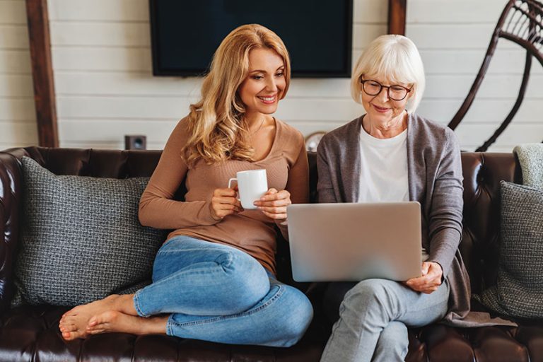 Smiling senior woman and her middle aged daughter sitting relax on sofa and drinking coffee in living room watching video on laptop together