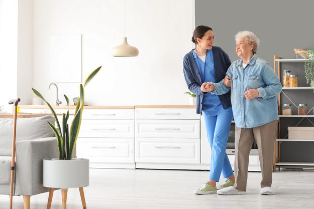Young caregiver helping senior woman to walk in kitchen — Photo