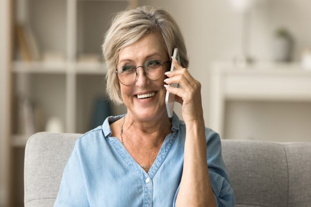 Cheerful pretty senior woman in stylish eyeglasses enjoying telephone call Cheerful pretty blonde senior woman in stylish eyeglasses enjoying telephone call, connection, funny conversation on home sofa, looking away with toothy smile, listening, speaking, laughing — Photo