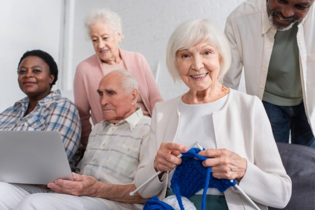 Elderly woman holding yarn near multiethnic friends with laptop in nursing home — Photo