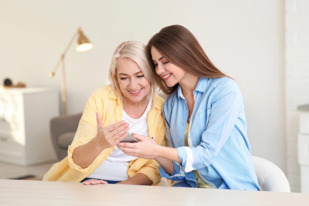 Portrait of young woman and her mature mother with mobile phone at table indoors — Photo