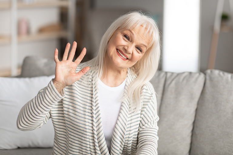 elderly-woman-waving-at-camera-and-smiling-sitting-on-sofa