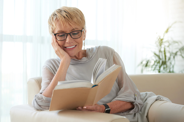 senior-blond-woman-glasses-resting-sofa-reading-book
