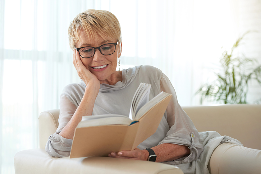 senior-blond-woman-glasses-resting-sofa-reading-book