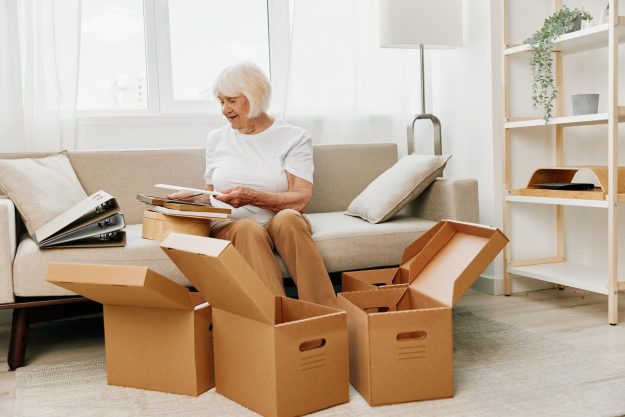 elderly woman sits on a sofa at home with boxes. collecting things with memories albums with photos and photo frames moving to a new place cleaning things and a happy smile. Lifestyle retirement.
