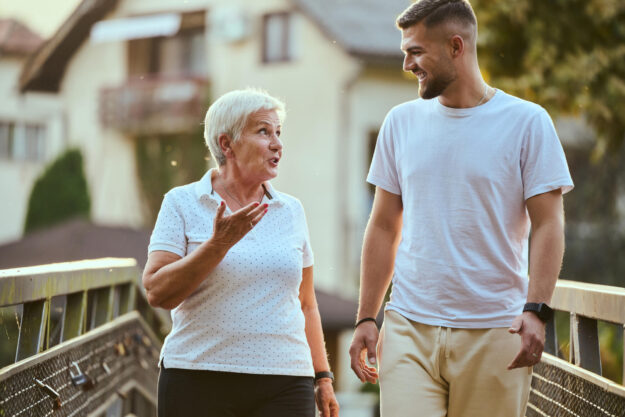 A handsome man and an older woman share a serene walk in nature, crossing a beautiful bridge against the backdrop of a stunning sunset, embodying the concept of a healthy and vibrant intergenerational life.